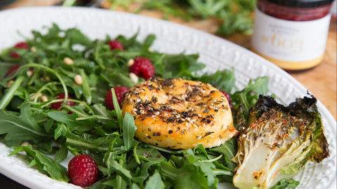 Kräuter-Grillkäse auf Rucolasalat mit Pinienkernen, Himbeeren und Honig auf weißem Teller.