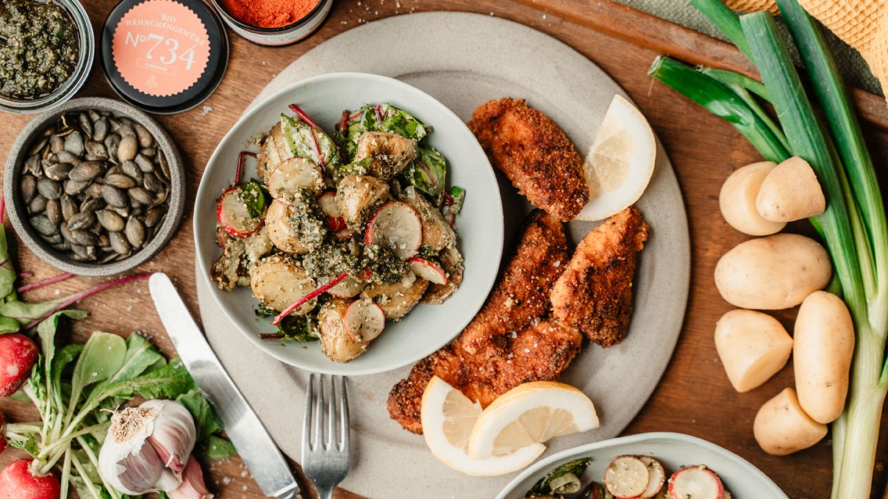 Ein Teller mit panierten Hähnchenstreifen, Kartoffelsalat, Gemüse und Zitronenschnitzen.