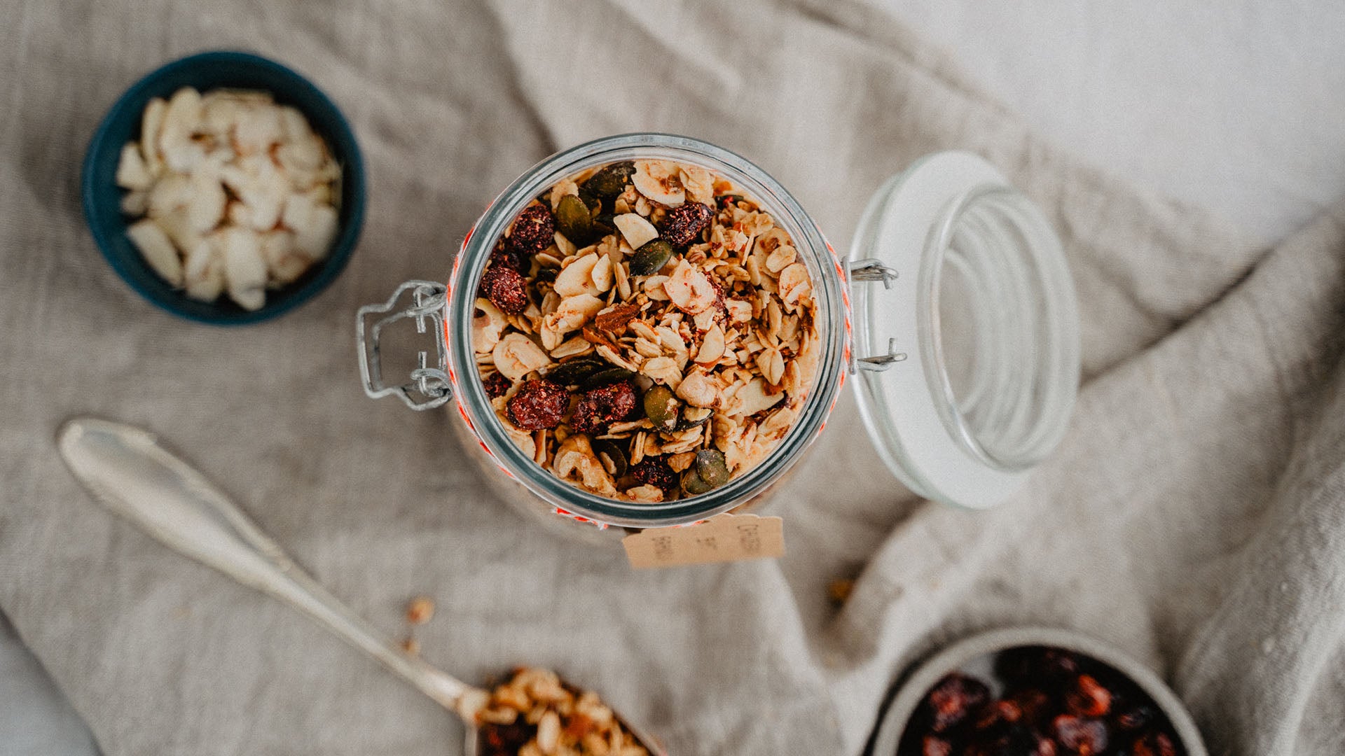 Ein Glas Müsli mit Kernen und Trockenfrüchten, daneben Schälchen mit Mandelblättchen und Cranberries.