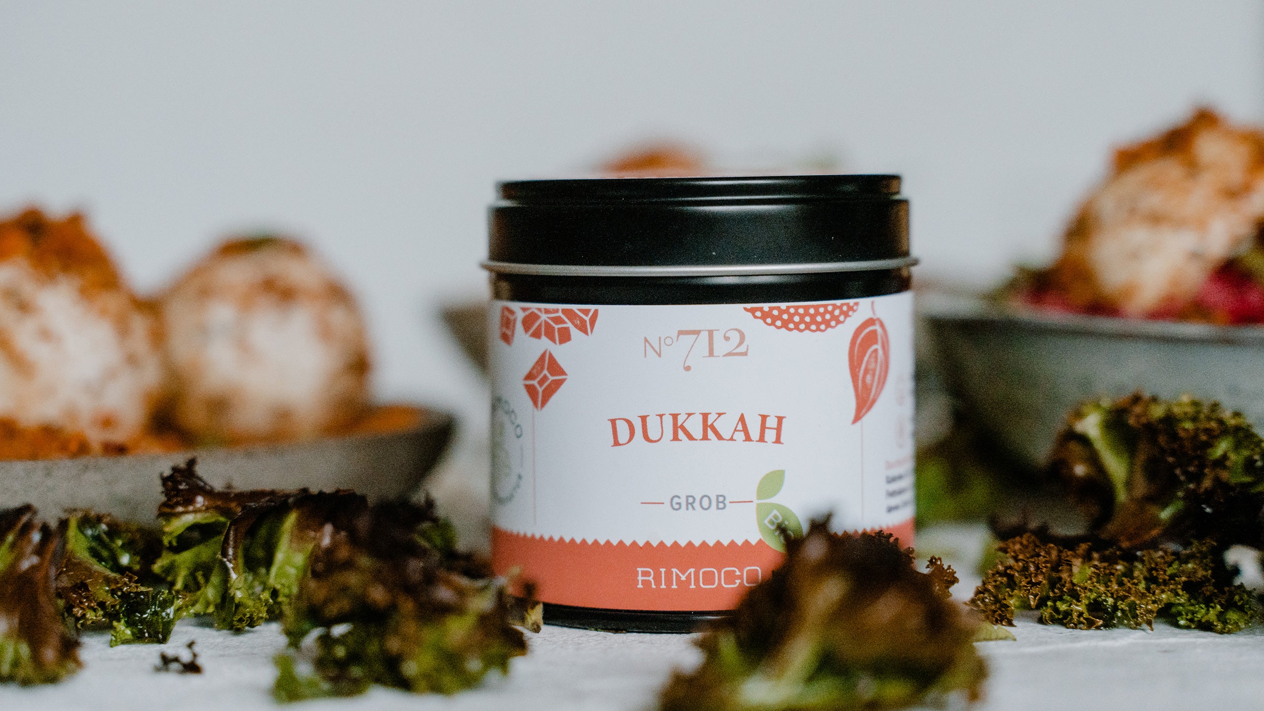 Ein Glas mit Dukkah-Gewürzmischung, dahinter Grünkohlchips und bunte Schüsseln mit Essen.