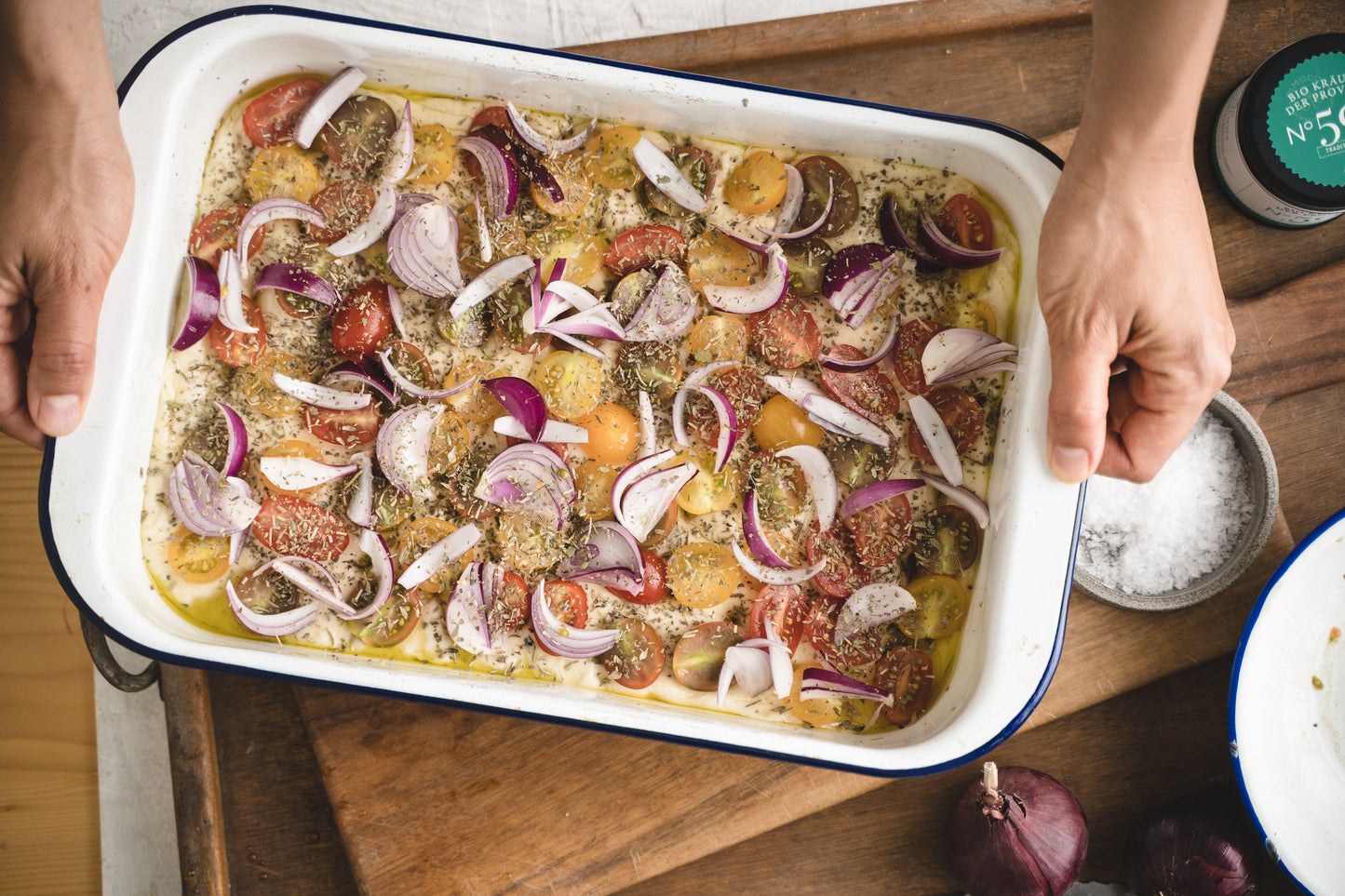 Hände halten eine Auflaufform mit Tomaten, roten Zwiebeln, grobem Gewürz und Olivenöl.