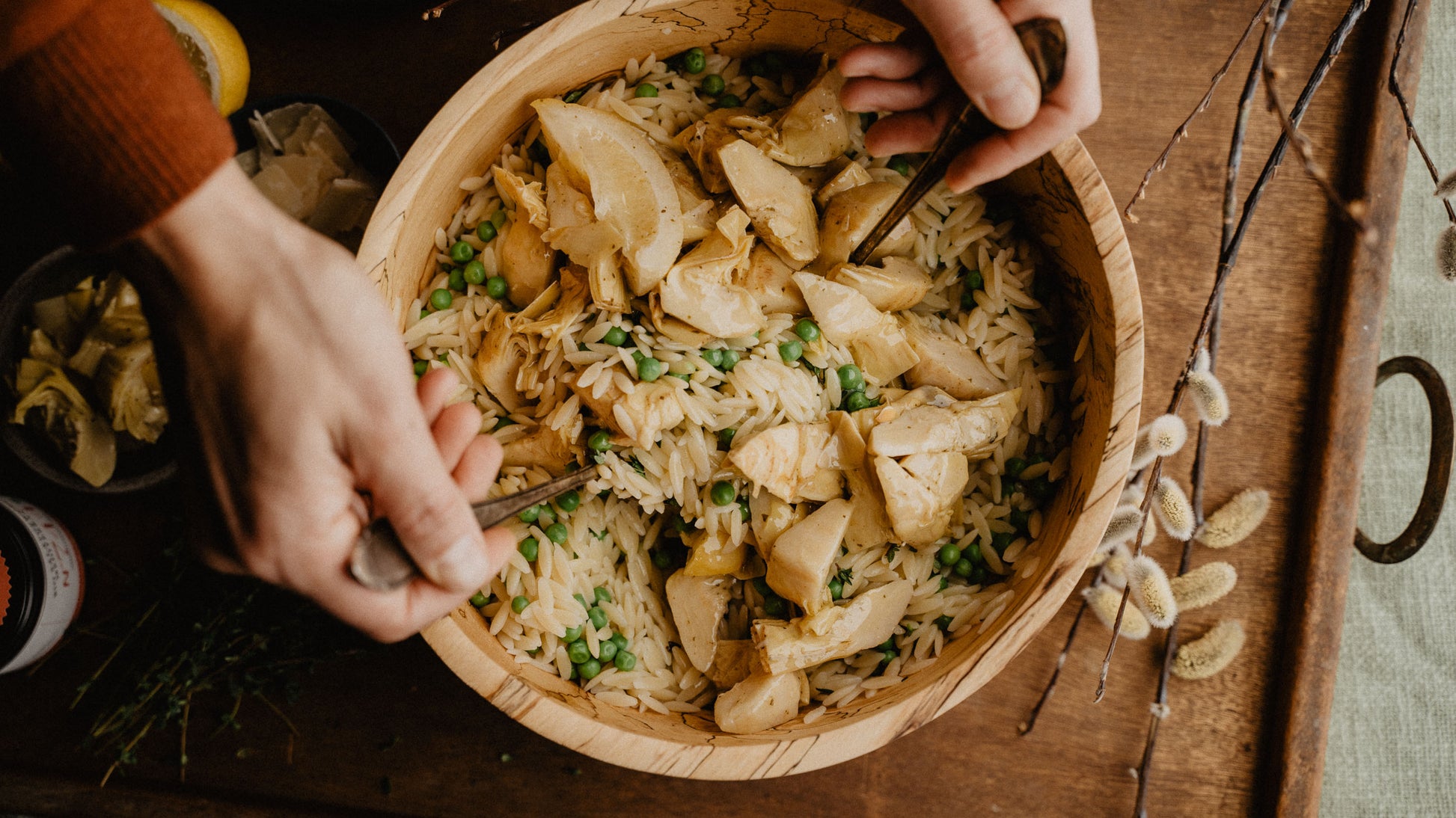 Hände mischen Orzo-Nudeln mit Erbsen, Artischocken und sichtbaren Gewürzmischungen in Holzschüssel.