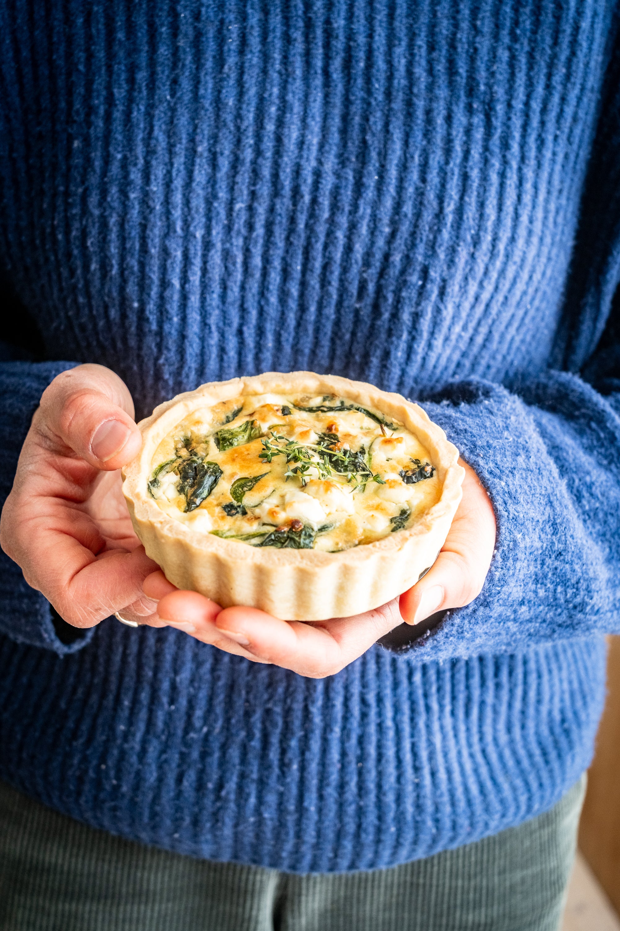 Eine Person in einem blauen Pullover hält mit beiden Händen eine Quiche mit Käse und grünem Gemüse in der Hand.