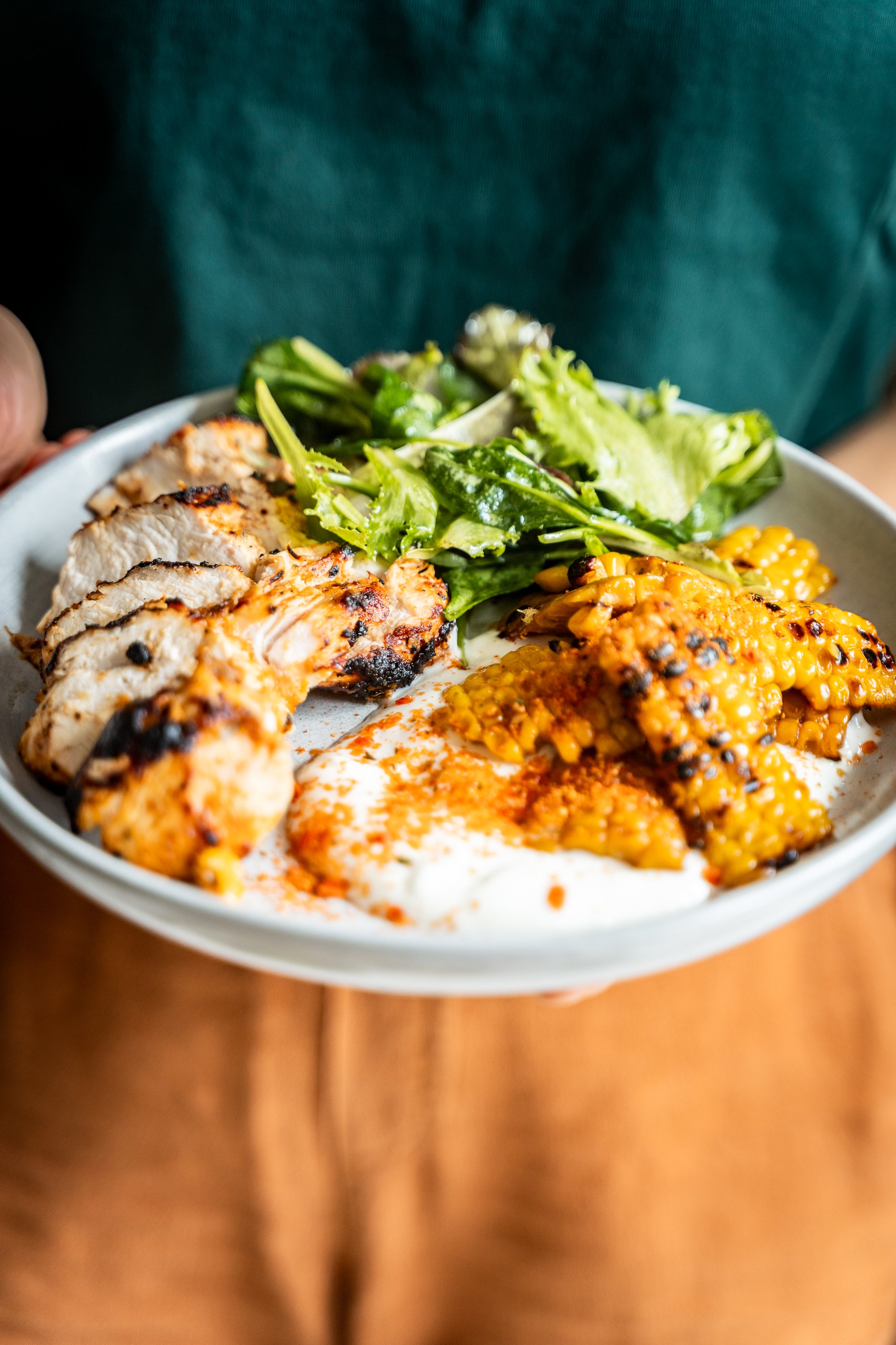 Ein Teller mit gegrilltem Hähnchen, Maiskolben, Salatblättern und weißem Dressing.