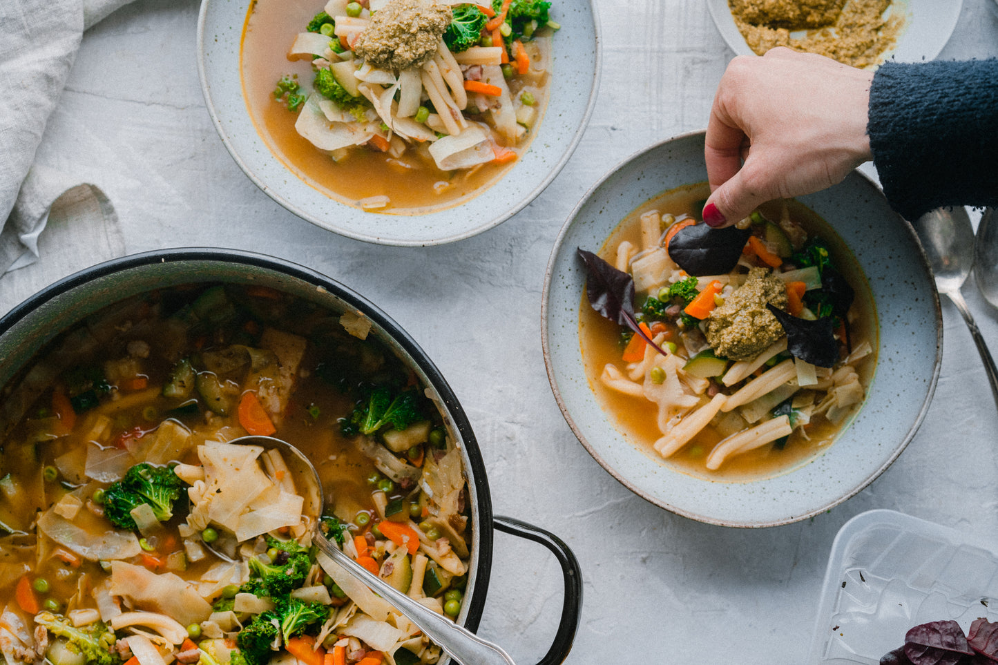 Eine Hand garniert Schüsseln Gemüsenudelsuppe, daneben liegt Gewürzseminar Italien auf dem Tisch.
