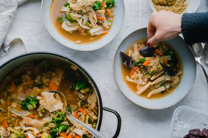 Eine Hand garniert Schüsseln Gemüsenudelsuppe, daneben liegt Gewürzseminar Italien auf dem Tisch.