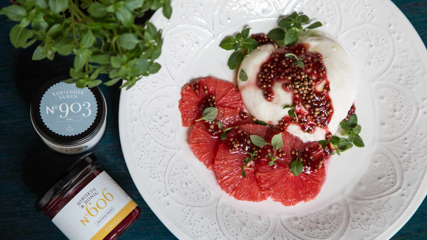 Teller mit Burrata, Grapefruit, Microgreens, roter Sauce und Gläsern Hibiskus & Honig daneben.