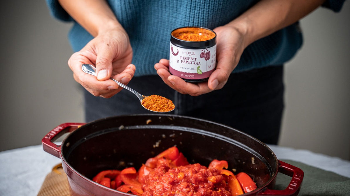 Tomaten und Paprika im Topf, eine Person streut mit einem Löffel rote Gewürze aus einem Glas hinein.