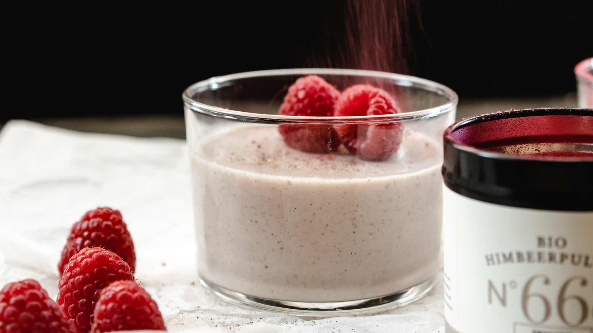 Ein Glas mit cremigem Dessert und frischen Himbeeren, daneben weitere Beeren und ein Behälter.