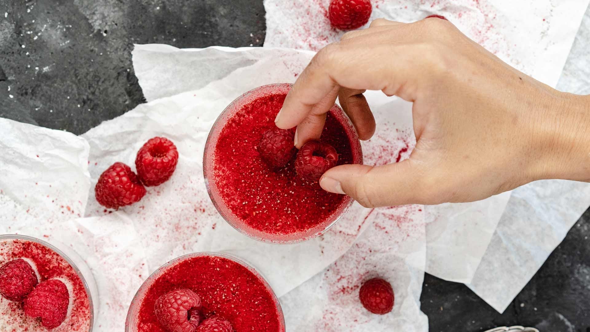 Eine Hand legt eine Himbeere auf ein rotes Dessert, daneben liegen frische Himbeeren auf Backpapier.