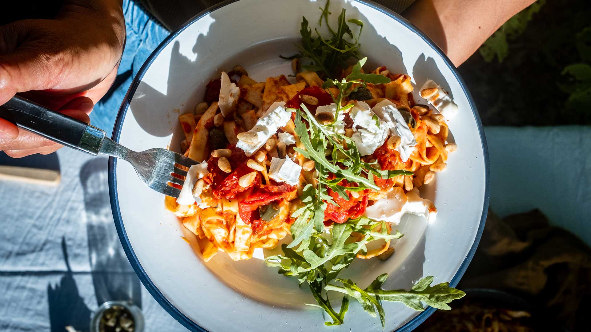Eine Hand hält eine Schüssel Pasta mit Tomatensoße, Rucola, Käse, Pinienkernen und Kräutern.