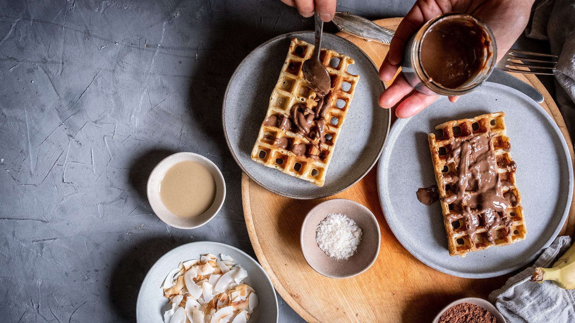 Waffeln auf Tellern, eine davon mit Schokolade überzogen, umgeben von Schälchen mit Toppings.