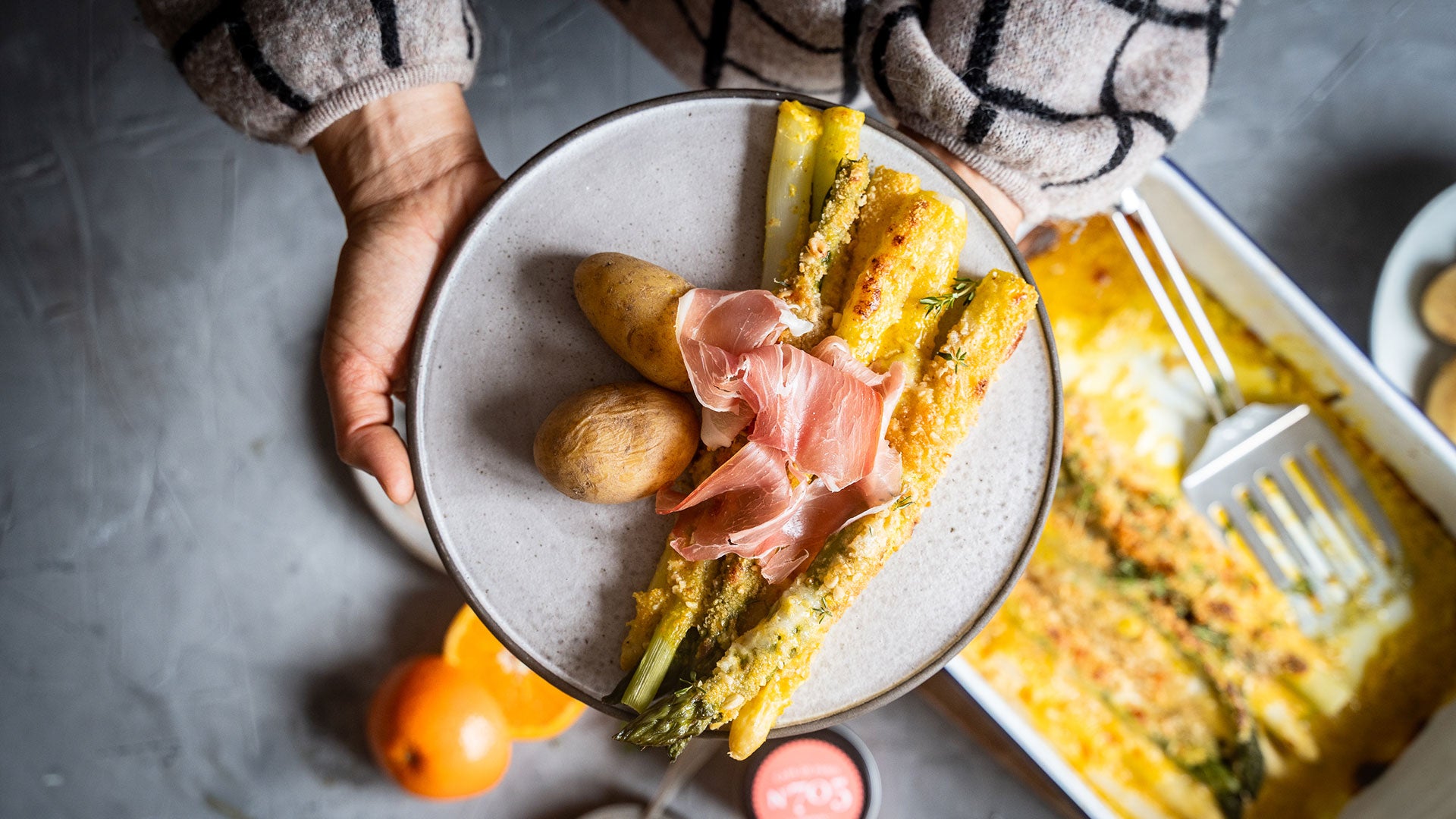 Ein Teller mit Spargel, Kartoffeln und Prosciutto-Scheiben wird über einer grauen Tischplatte gehalten.